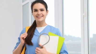 Smiling nurse with backpack holding papers, exploring PhD in Nursing and DNP degree options.